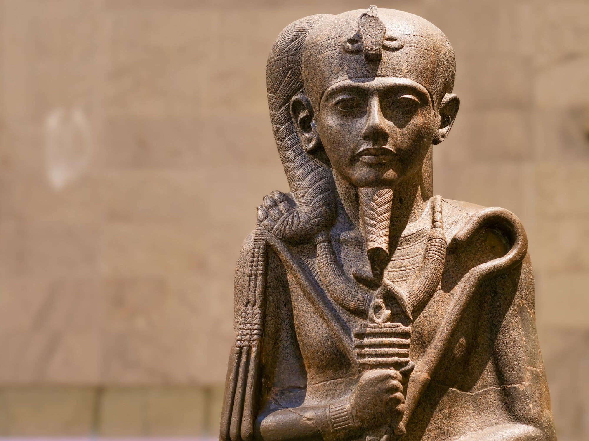 Close-up of an ancient Egyptian stone statue of the god Khonsu as a child, with a braided sidelock of youth, uraeus on the forehead, and regalia including a crook, flail, and composite scepter.